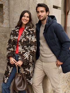 Eine Frau in einem gemusterten Pelzmantel und einem roten Oberteil steht lächelnd neben einem Mann in einer marineblauen Winterjacke und einem beigen Pullover. Sie stehen draußen an einer Steinmauer, und die Frau hält eine braune Handtasche in der Hand.