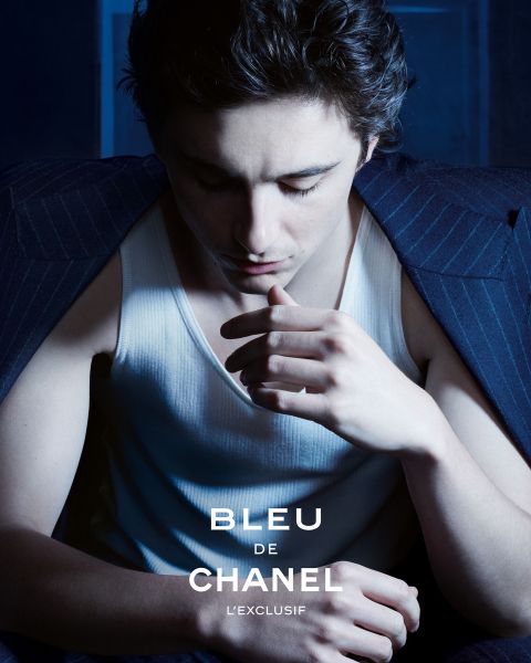 Ein junger Mann in Nadelstreifenanzugjacke und weißem Tank-Top blickt nachdenklich nach unten, beleuchtet von sanftem blauem Licht. Der Text "BLEU DE CHANEL L'EXCLUSIF" ist am unteren Rand des Bildes eingeblendet.