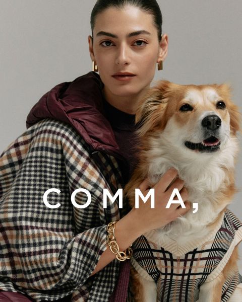 Eine Frau in einem karierten Mantel und goldenen Accessoires posiert mit einem hellbraunen und weißen Hund in einem passenden karierten Outfit. Das Wort "COMMA" erscheint in großer weißer Schrift quer über das Bild.
