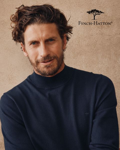 Ein Mann mit gewelltem braunem Haar und Bart trägt einen marineblauen Pullover und steht vor einer beigefarbenen, strukturierten Wand. Das Logo und der Name von Fynch-Hatton erscheinen in der oberen rechten Ecke.