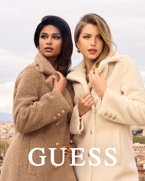 Zwei Frauen stehen dicht beieinander im Freien und tragen strukturierte Mäntel, eine in hellbraun und eine in cremefarben. Beide halten ihren Kragen fest und schauen nach vorne. Das Wort "GUESS" steht in fetten weißen Buchstaben am unteren Rand.