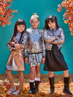 Drei junge Mädchen stehen selbstbewusst zwischen Herbstblättern und tragen metallisch glänzende Outfits. Zwei Mädchen tragen silberne Jacken, und das Mädchen in der Mitte trägt ein mit Pailletten besetztes Oberteil und einen Rock. Ein blaugrüner Hintergrund kontrastiert mit orangefarbenem Herbstlaub.