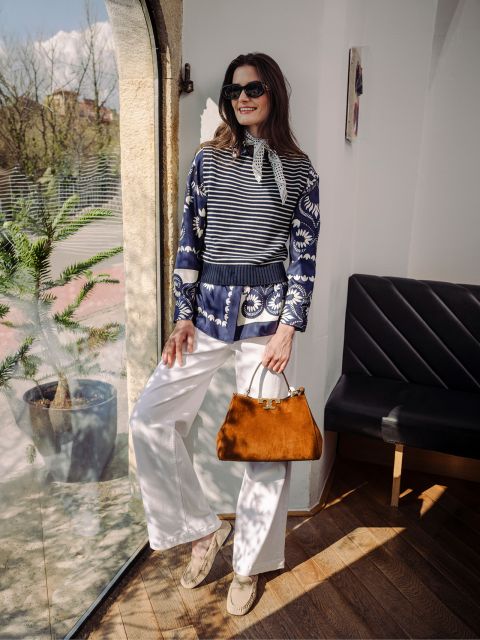 Eine Frau mit Sonnenbrille, blau gemustertem Oberteil und weißer Hose steht an einem Fenster, hält eine braune Handtasche in der Hand und lächelt.