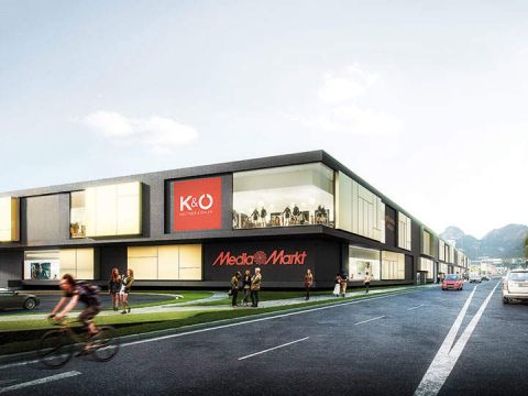 Modernes Geschäftsgebäude mit Media Markt- und K&Ö-Schildern, Menschen, die auf dem Bürgersteig gehen, und ein Radfahrer, der auf der Straße vorbeifährt.