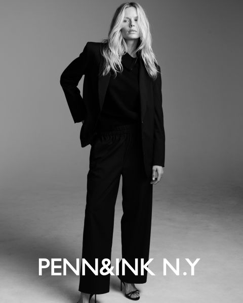 Eine Frau mit langem, gewelltem Haar trägt einen dunklen Anzug und hochhackige Sandalen und steht selbstbewusst in einem Studio mit einfarbigem Hintergrund. Der Text am unteren Rand lautet "PENN&INK N.Y.