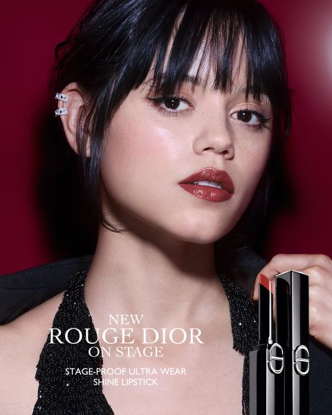 Eine Frau mit glänzendem dunkelrotem Lippenstift posiert vor einem tiefroten Hintergrund und trägt ein glitzerndes schwarzes Neckholder-Top und Ohrstulpen. Sie hält eine Tube des Rouge Dior Shine Lipstick in der Hand. Der Text wirbt für den neuen bühnenfesten Ultra-Wear-Lippenstift.