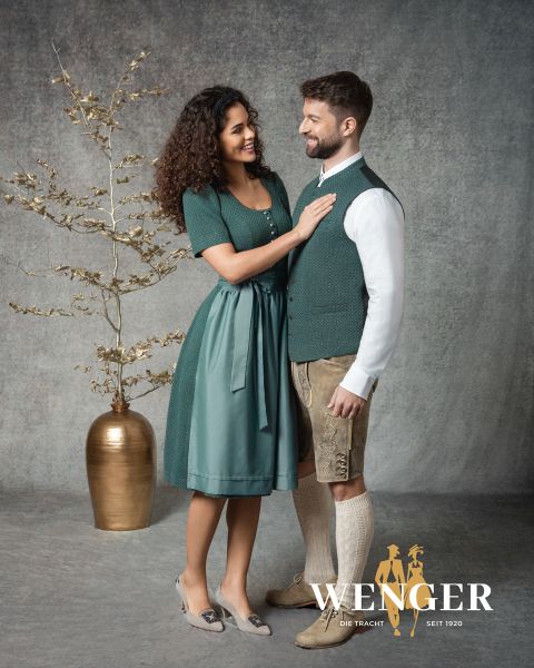 Eine lächelnde Frau in einem grünen Dirndl steht neben einem Mann in traditioneller bayerischer Kleidung, darunter Lederhosen und eine Weste. Sie stehen sich vor einem Studiohintergrund mit einer goldenen Vase und einem dekorativen Zweig gegenüber. "Wenger"-Logo am unteren Rand.