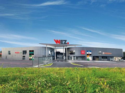 Ein modernes Einkaufszentrum mit einem großen "WEZ"-Schild auf dem Dach, einer grauen Fassade, verschiedenen Ladenschildern und einem klaren blauen Himmel darüber.