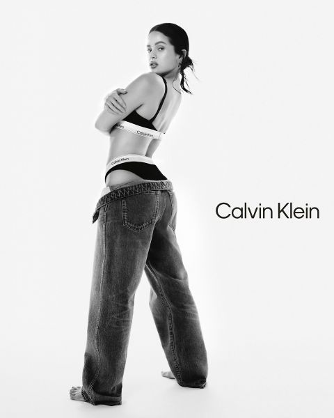 Eine Frau posiert in Calvin Klein Unterwäsche und weiten Jeans und schaut über ihre Schulter. Der Hintergrund ist einfarbig weiß mit dem Calvin Klein-Logo in schwarzer Schrift auf der rechten Seite. Das Bild ist in Schwarz-Weiß gehalten.