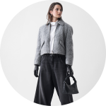 Eine Frau, die eine graue wattierte Jacke, eine schwarze Jeans mit weitem Bein, ein weißes Oberteil, schwarze Handschuhe und eine kleine schwarze Handtasche trägt, steht vor einem schlichten weißen Hintergrund und blickt nach links.