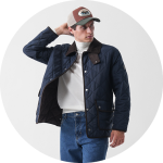 Ein Mann, der eine marineblaue Steppjacke, einen weißen Rollkragenpullover, blaue Jeans und eine braun-beigefarbene Baseballmütze trägt, steht mit einer Hand hinter dem Kopf vor einem schlichten weißen Hintergrund.