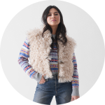 Eine Frau steht selbstbewusst in einer flauschigen beigen Weste über einem bunt gemusterten Pullover und blauen Jeans, die Hand in der Tasche, vor einem schlichten weißen Hintergrund.