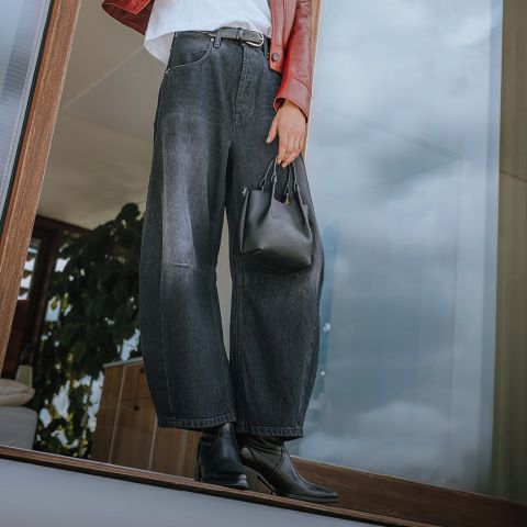 Person, die in einem Innenraum in der Nähe eines Fensters steht. Sie trägt eine weite schwarze Jeans, schwarze Ankle Boots, eine rote Lederjacke und hält eine kleine schwarze Handtasche in der Hand. Das Foto ist aus einem niedrigen Winkel aufgenommen, wobei der Fokus auf dem Outfit liegt.
