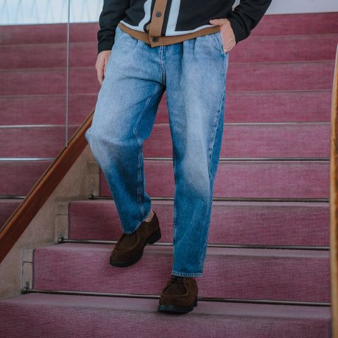 Eine Person, die weite blaue Jeans, eine dunkle Jacke und braune Schuhe trägt, steht auf einer Treppe mit rosa Teppichboden und hat eine Hand in der Tasche. Das Gesicht ist nicht zu erkennen.