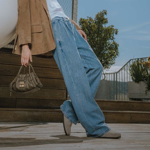 Eine Person, die weite blaue Jeans, einen hellbraunen Blazer und beigefarbene Schuhe trägt, steht im Freien und hält eine kleine olivgrüne Handtasche. Im Hintergrund sind Holzstufen, Pflanzen und ein Metallgeländer vor einem blauen Himmel zu sehen.