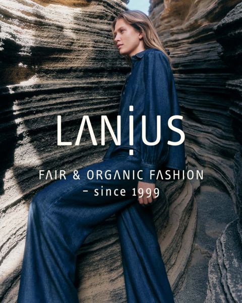 Eine Frau in einem langen, dunkelblauen Kleid sitzt zwischen geschichteten Felsformationen. Der überlagerte Text lautet: "LANIUS FAIR & ORGANIC FASHION - seit 1999.