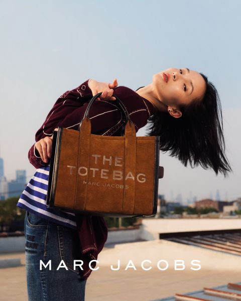 Eine Frau posiert im Freien mit einer braunen Marc-Jacobs-Tasche in der Hand, lehnt sich mit geneigtem Kopf zurück und trägt eine kastanienbraune Jacke und blaue Jeans. Auf dem Bild erscheinen die Worte "THE TOTE BAG MARC JACOBS" und "MARC JACOBS".