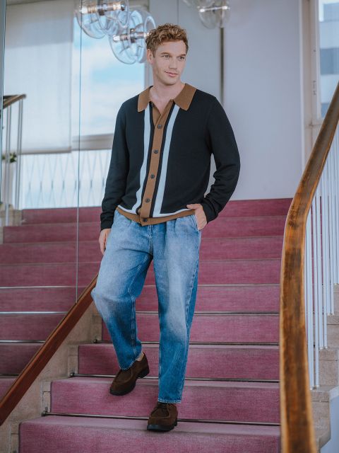 Ein Mann mit hellem Haar geht eine Treppe mit rosa Teppich hinunter. Er trägt eine schwarze Strickjacke mit hellbraunen und grauen Streifen, blaue Jeans und braune Schuhe. Der Schauplatz scheint ein Innenraum mit moderner Einrichtung zu sein.