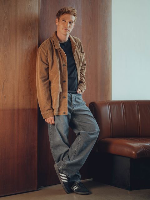 Ein Mann mit kurzen hellbraunen Haaren steht lässig an einer holzgetäfelten Wand. Er trägt eine braune Jacke, ein schwarzes Hemd, graue weite Jeans und schwarze Adidas-Turnschuhe, eine Hand in der Tasche und einen entspannten Gesichtsausdruck.
