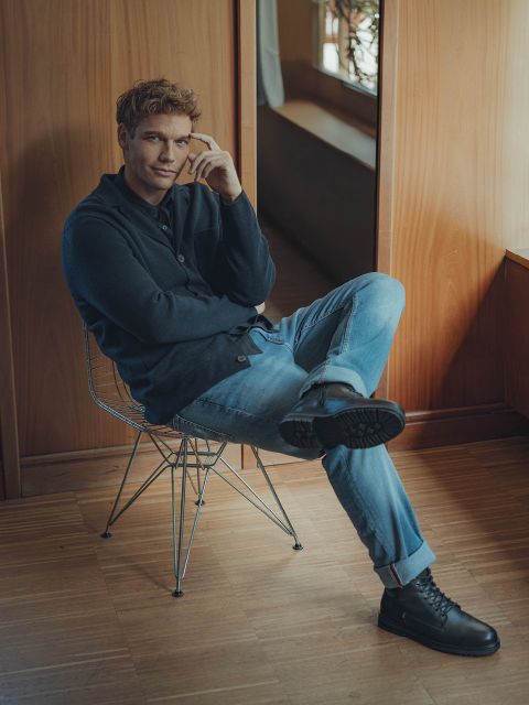 Ein Mann mit hellbraunem Haar, der einen dunklen Pullover, blaue Jeans und schwarze Schuhe trägt, sitzt lässig auf einem Drahtstuhl mit übereinander geschlagenen Beinen in einem holzgetäfelten Raum und lächelt leicht in die Kamera.