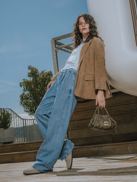 Eine Frau mit gewelltem Haar steht im Freien auf einer Holztreppe. Sie trägt einen hellbraunen Blazer, ein weißes Hemd, Jeans mit weitem Bein und beigefarbene Schuhe. Sie hält eine kleine olivgrüne Handtasche in der Hand und blickt selbstbewusst in die Kamera. Im Hintergrund ist der blaue Himmel zu sehen.