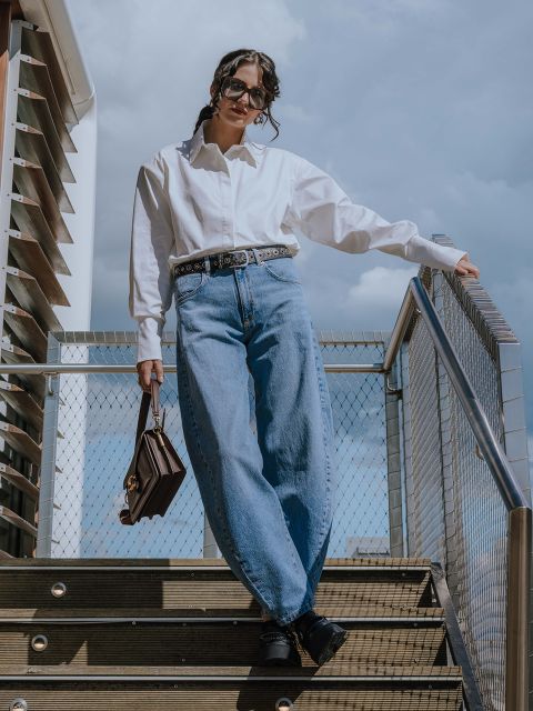 Eine Person, die ein weißes Button-up-Hemd, blaue Jeans mit hoher Taille, schwarze Schuhe und eine Sonnenbrille trägt, steht auf einer Außentreppe, hält eine braune Handtasche in der Hand und stützt sich mit einer Hand auf ein Geländer vor einem bewölkten Hintergrund.
