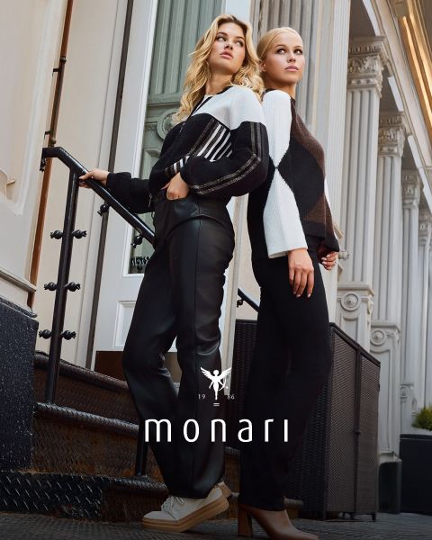 Zwei Frauen stehen auf einer Freitreppe vor einem Gebäude mit Säulen, beide tragen modische schwarz-weiße Pullover und schwarze Hosen. Das "monari"-Logo ist am unteren Rand des Bildes zu sehen.