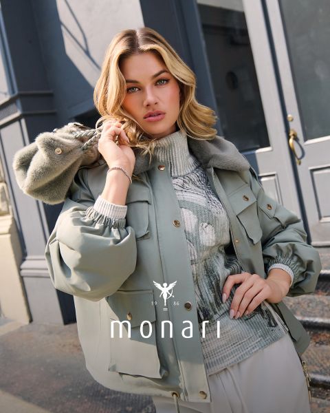 Eine Frau mit blondem Haar posiert im Freien in einer eleganten hellgrauen Jacke über einem gemusterten Pullover und hält den Kunstpelzkragen der Jacke. Der Hintergrund besteht aus großen Fenstern und grauen Gebäudefassaden. Am unteren Rand steht "Monari" geschrieben.