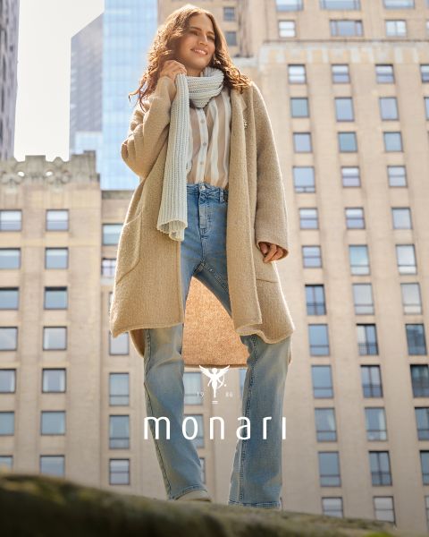 Eine lächelnde Frau, die einen beigen Mantel, ein gestreiftes Hemd, blaue Jeans und einen hellen Schal trägt, steht im Freien vor einem hohen Gebäude. Das Monari-Logo ist am unteren Rand des Bildes zu sehen.