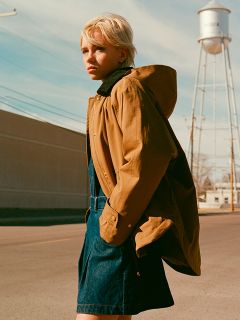 Eine Person mit kurzem blondem Haar steht auf einer Straße und trägt eine braune Jacke über einem blauen Kleid. Im Hintergrund sind bei klarem Himmel ein Wasserturm und Stromleitungen zu sehen.
