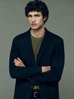 Ein Mann mit lockigem dunklem Haar, der einen schwarzen Blazer über einem grünen Rippstrickpullover trägt, steht mit verschränkten Armen vor einem einfarbigen grauen Hintergrund und blickt mit neutraler Miene geradeaus.