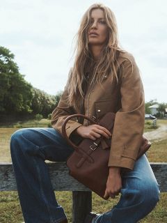 Eine Frau mit langen blonden Haaren sitzt im Freien auf einem Holzzaun, trägt eine hellbraune Jacke, blaue Jeans und hält eine braune Handtasche in ihrem Schoß. Im Hintergrund sind Bäume und ein Weg zu sehen.