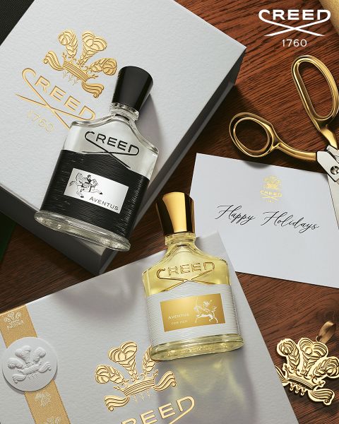 Zwei Creed-Parfümflakons (Aventus und Aventus for Her) werden in eleganten weißen Geschenkschachteln mit goldenen Logos zusammen mit einer goldenen Schere und einer "Happy Holidays"-Karte auf einem Holztisch präsentiert.