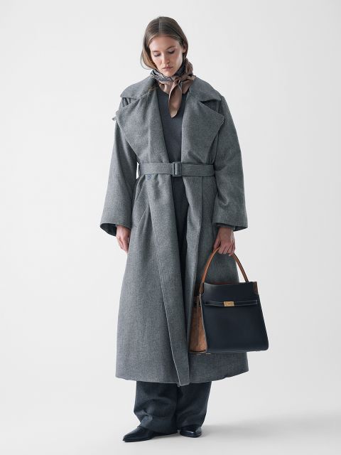 Eine Frau steht vor einem weißen Hintergrund und trägt einen langen grauen Mantel mit Gürtel, eine graue Hose und schwarze Schuhe. Sie hat einen braunen Schal um den Hals und hält eine schwarze Handtasche mit braunen Griffen in der linken Hand.