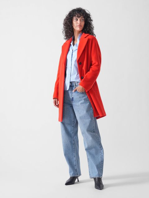 Eine Frau mit lockigem Haar trägt einen leuchtend roten Mantel über einem hellblauen Hemd, kombiniert mit einer locker sitzenden Bluejeans und schwarzen Stiefeln mit Absätzen, die vor einem schlichten weißen Hintergrund stehen.