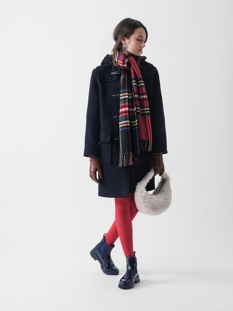 Eine Frau in schwarzem Mantel, roter Strumpfhose und marineblauen Stiefeln, die einen gestreiften Schal und braune Handschuhe trägt, hält eine flauschige weiße Handtasche. Sie steht vor einem schlichten weißen Hintergrund und blickt zur Seite.