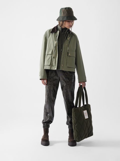 Eine Person, die eine grüne Jacke, eine dunkle, bedruckte Hose, braune Stiefel und einen grünen Hut trägt und eine gesteppte grüne Tragetasche in der Hand hält, steht vor einem schlichten weißen Hintergrund und blickt nach unten und zur Seite.