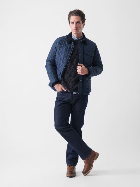 Ein Mann steht vor einem schlichten weißen Hintergrund und trägt eine dunkelblaue Steppjacke, einen schwarzen Pullover, ein weißes Hemd mit Kragen, dunkelblaue Jeans und braune Stiefel. Er kreuzt ein Bein und hat eine Hand in der Tasche.
