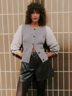 Eine Frau mit lockigem Haar steht an einer gekachelten Wand. Sie trägt eine hellgraue, wuschelige Strickjacke, schwarze, hoch sitzende Ledershorts und eine schwarze Strumpfhose, die Hände in den Taschen und einen selbstbewussten Gesichtsausdruck.