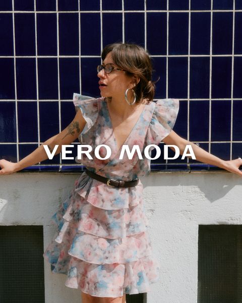 Eine Frau mit Brille und einem gerüschten Blumenkleid lehnt an einer weißen Wand vor einem blau gekachelten Hintergrund. Der Schriftzug "VERO MODA" erscheint in der Mitte des Bildes.