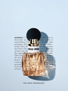Ein Flakon von Miu Miu-Parfüm mit einer runden schwarzen Kappe befindet sich vor einem verschwommenen Text auf weißem Hintergrund. Das Parfüm ist hell bernsteinfarben, und der Text darunter lautet "THE NEW FRAGRANCE.
