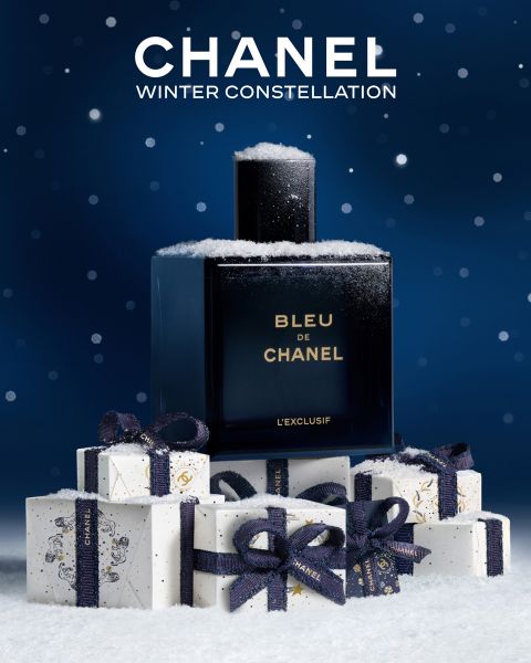 Ein mit Schnee bestäubter Flakon des Parfums Bleu de Chanel steht auf gestapelten, verschneiten Geschenkschachteln, die mit marineblauen Bändern verschnürt sind, vor einem sternblauen Hintergrund mit dem Text "CHANEL WINTER CONSTELLATION" darüber.