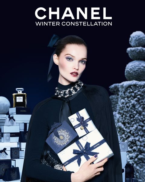 Eine Frau in eleganter schwarzer Kleidung hält gestapelte Chanel-Geschenkschachteln, umgeben von weiteren Geschenkschachteln und einer Chanel-Parfümflasche, über der auf dunklem Hintergrund "CHANEL WINTER CONSTELLATION" steht.
