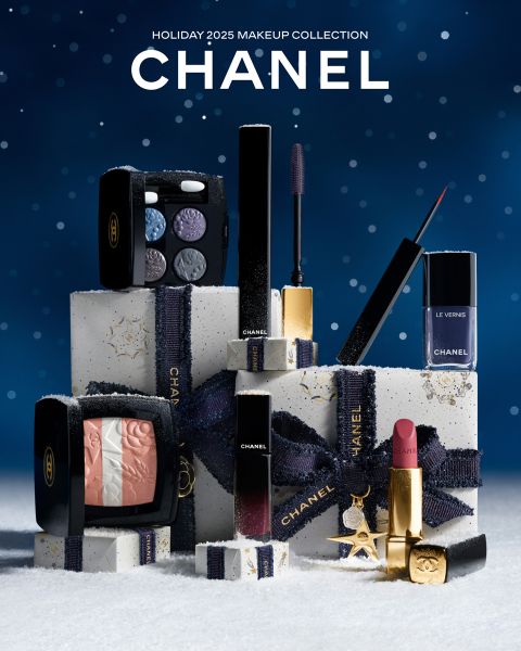 Chanel Holiday 2025 Make-up-Kollektion Display mit Lippenstiften, Lidschatten-Palette, Nagellack, Mascara und erröten angeordnet auf weißen Geschenk-Boxen mit marineblauem Band gebunden, vor einem verschneiten, blauen Hintergrund gesetzt.
