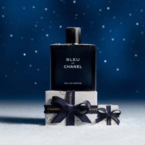 Ein Flakon des Parfums Bleu de Chanel steht auf verschneitem Boden, umgeben von kleinen, weißen Geschenkschachteln, die mit dunkelblauen Bändern verschnürt sind. Schneeflocken fallen sanft vor einem dunkelblauen, festlichen Hintergrund.