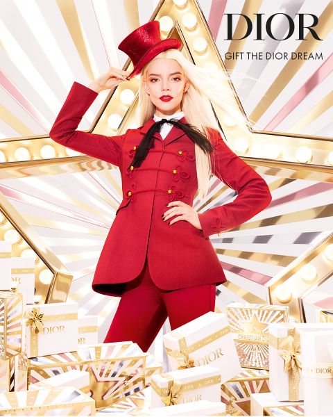 Eine Frau in einem auffälligen roten Anzug und Zylinder posiert selbstbewusst vor einem großen beleuchteten Stern, umgeben von weißen und goldenen Dior-Geschenkboxen. Der Text lautet "DIOR GIFT THE DIOR DREAM.