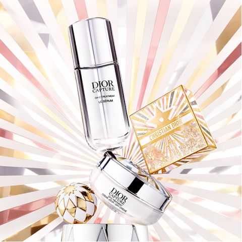 Zwei Dior-Pflegeprodukte und eine goldene Christian-Dior-Schminkdose werden auf einem strahlenden Hintergrund mit Sonnenschliffmustern in Gold-, Rosa- und Weißtönen präsentiert, die einen luxuriösen und leuchtenden Effekt erzeugen.