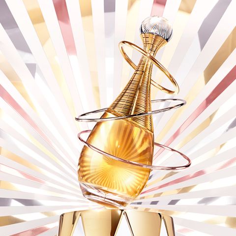 Ein goldener Parfümflakon mit eleganten Spiralringen steht vor einem strahlenden Hintergrund aus goldenen, rosafarbenen und weißen Strahlen, der ein luxuriöses und glamouröses Erscheinungsbild erzeugt.