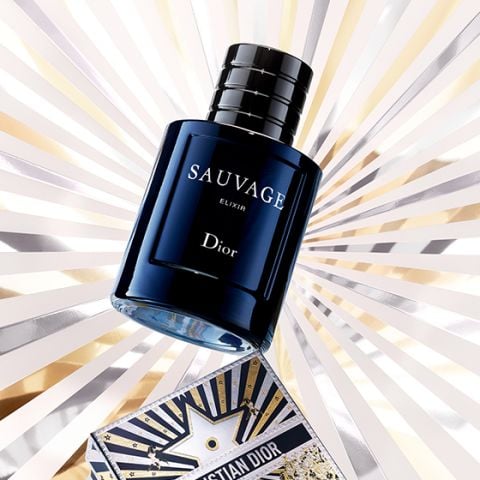Ein dunkelblauer Flakon des Parfums Dior Sauvage Elixir ist vor einem leuchtenden Hintergrund mit Sternenhimmel ausgestellt, wobei eine dekorative Karte von Christian Dior am unteren Rand teilweise sichtbar ist.