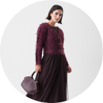 Eine Frau, die eine strukturierte kastanienbraune Jacke und einen langen, fließenden kastanienbraunen Rock trägt, hält eine passende Handtasche. Sie steht vor einem schlichten weißen Hintergrund und blickt leicht zur Seite.